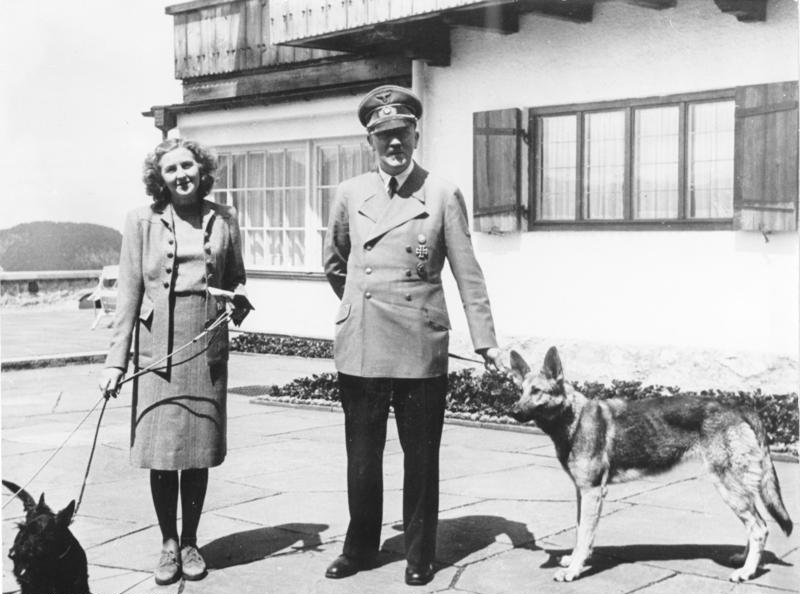 Eva Braun and Adolf Hitler standing outside a building with their two dogs